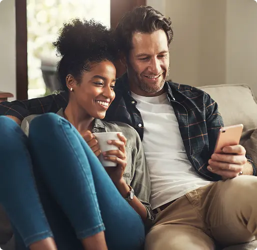 Couple sitting on a couch, smiling while looking at a smartphone together at home